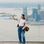 A person standing on a high observation deck, taking a selfie with a city skyline and water in the background.