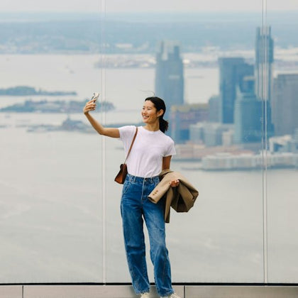 A person standing on a high observation deck, taking a selfie with a city skyline and water in the background.
