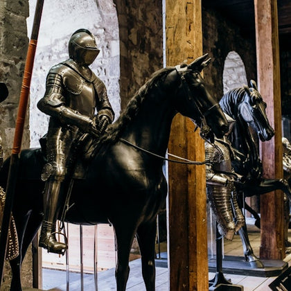 Two armored knight mannequins on horseback in a dimly lit stone-walled room with large arched windows.