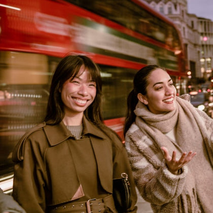 Two women smiling and chatting on a city street with a blurred red double-decker bus passing by in the background.