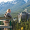 A woman with a braid stands on a wooden platform overlooking a valley with dense trees and a large historic building, backed by mountains.