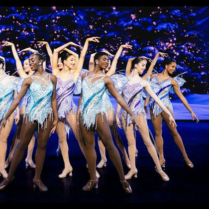 A group of dancers in sparkly costumes performing on a stage with a backdrop of a snowy, starry scene.