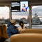 Passengers seated inside a ferry with a view of a warship and cityscape through large windows, and a screen displaying a message.