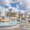 A grand neoclassical building with columns and a dome, surrounded by a square with fountains and visitors walking.