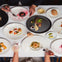 A top-down view of a table set with gourmet dishes, wine glasses, and bread. Two people are visible with utensils.