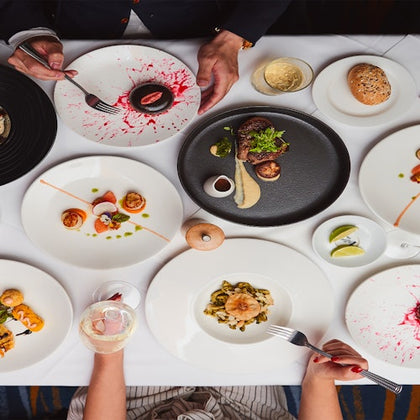 A top-down view of a table set with gourmet dishes, wine glasses, and bread. Two people are visible with utensils.