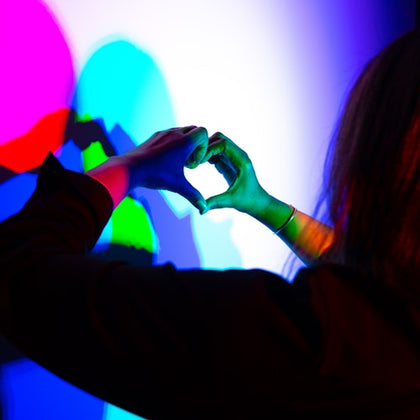 A person forming a heart shape with their hands in front of a colorful light projection creating overlapping shadows.