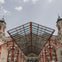 Symmetrical view of a historic building's entrance with a large, red-framed glass canopy and towers on either side under a partly cloudy sky.