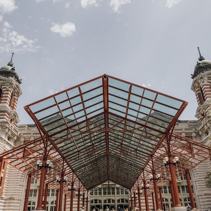 Symmetrical view of a historic building's entrance with a large, red-framed glass canopy and towers on either side under a partly cloudy sky.