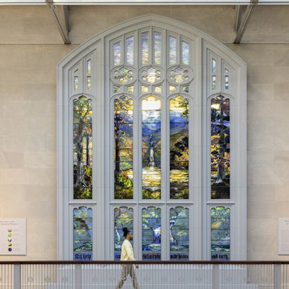 A person walks past a large, colorful stained glass window depicting a natural landscape inside a building.