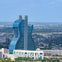 A large blue glass skyscraper shaped like a guitar, set in a cityscape with greenery and buildings in the background.