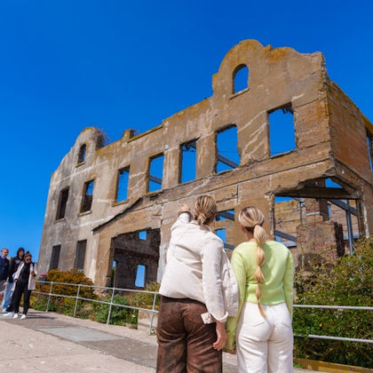 Two people in the foreground pointing at a ruined building under a blue sky, with others observing in the background.
