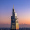 A modern skyscraper with reflective glass and a spire at sunset, against a gradient sky and cityscape in the background.