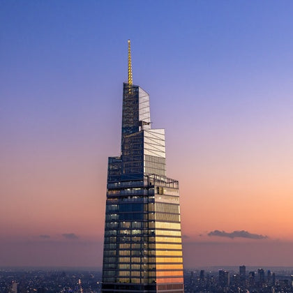 A modern skyscraper with reflective glass and a spire at sunset, against a gradient sky and cityscape in the background.
