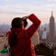 A person in a red coat takes a photo of a cityscape with skyscrapers, including the Empire State Building, at sunset.