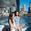 Two women standing on a boat with a city skyline and water in the background on a sunny day.
