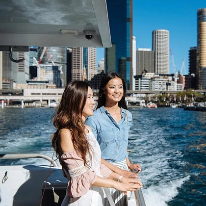 Two women standing on a boat with a city skyline and water in the background on a sunny day.