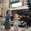 Two children interact with a large industrial wheel in a museum-like setting with machinery and a bright display in the background.