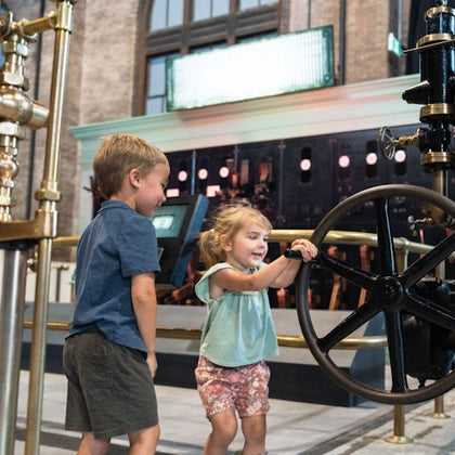 Two children interact with a large industrial wheel in a museum-like setting with machinery and a bright display in the background.