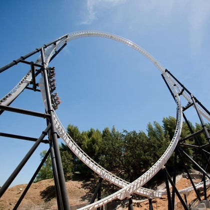A roller coaster with blue and green tracks, carrying passengers, moving on a wooden frame structure, surrounded by greenery.