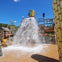 A water playground with a large bucket spilling water onto the ground, surrounded by wooden structures and slides under a clear sky.