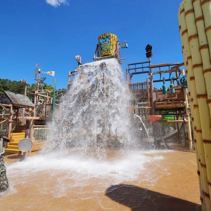 A water playground with a large bucket spilling water onto the ground, surrounded by wooden structures and slides under a clear sky.
