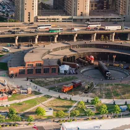 Aerial view of a roundhouse railway museum with trains, a turntable, pedestrians, and a highway in the background.