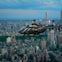 A black helicopter flies over a sprawling cityscape with dense buildings and skyscrapers, under a partly cloudy sky.