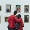Two people in red clothing stand closely together, looking at framed artworks displayed on a white gallery wall.
