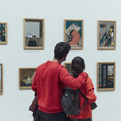Two people in red clothing stand closely together, looking at framed artworks displayed on a white gallery wall.