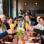 A group of smiling people sitting at a restaurant table, raising glasses filled with orange drinks in a toast.