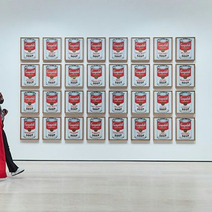 A gallery wall displaying 32 framed artworks of Campbell's soup cans in rows. A person in red walks by on the left.