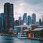 A waterfront cityscape with skyscrapers, a large Ferris wheel, and boats docked along a bustling pier under a cloudy sky.