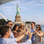 A group of people on a boat raising glasses in a toast with the Statue of Liberty visible in the background.