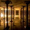A vast underground space with numerous evenly spaced columns and reflective water on the floor, illuminated by soft lighting.