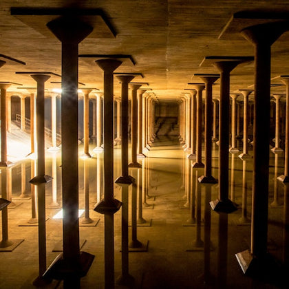A vast underground space with numerous evenly spaced columns and reflective water on the floor, illuminated by soft lighting.