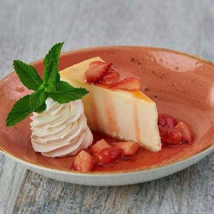 Slice of cheesecake with strawberry topping, a dollop of whipped cream garnished with mint, served on a terra cotta plate.