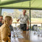 A man is speaking to a group seated around a table set with empty wine glasses in an outdoor venue with a scenic view.