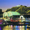An illuminated urban waterfront scene at dusk featuring a glass-domed building, colorful lights, and reflections on the water.