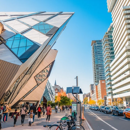 A modern, angular building beside a street with pedestrians, parked bicycles, and cars under a clear blue sky.