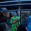 A man in a bright green costume plays a small red guitar on a bus with neon lighting, surrounded by onlookers.
