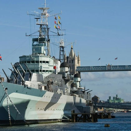 A historic battleship docked on a river with a bridge and tower in the background under a clear blue sky.