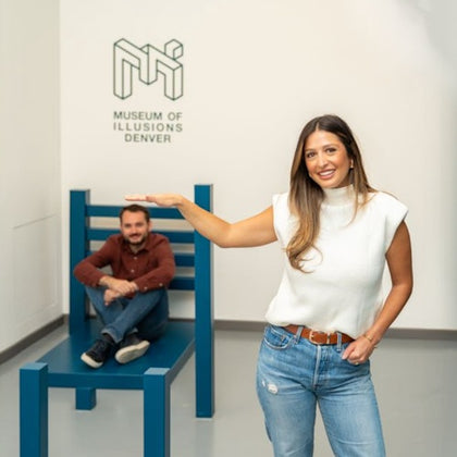 A woman poses in front of a large blue chair with a man sitting on it, creating an optical illusion at the Museum of Illusions Denver.