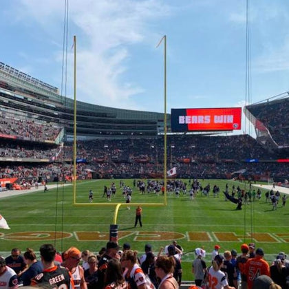 Crowd at a football game in a stadium, with "BEARS WIN" displayed on the scoreboard. Players are on the field.