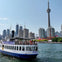 A sightseeing boat named 'Oriole' on water with the CN Tower and Toronto skyline in the background under a clear sky.