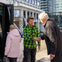 A man in a green checkered shirt shakes hands with another man near a vehicle, with a woman in a pink coat nearby.