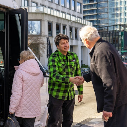 A man in a green checkered shirt shakes hands with another man near a vehicle, with a woman in a pink coat nearby.