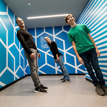 Three people lean at an angle in a blue room with geometric patterns at the Museum of Illusions in Boston.