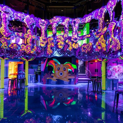 A vibrant, neon-lit interior with colorful abstract decor, a reflective floor, and art installations on the walls.