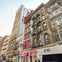 A city street with historic buildings, fire escapes, storefronts with pink awnings, and a bus partially visible in the foreground.